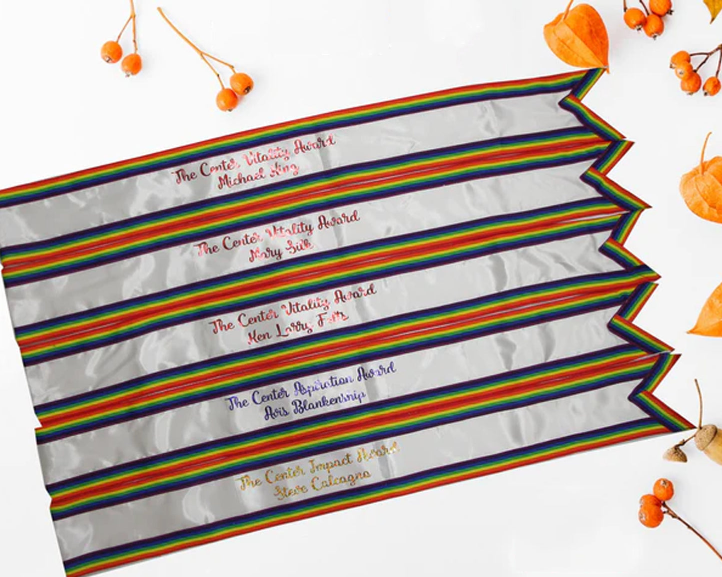 A set of silver award sashes with rainbow borders, embroidered with names and titles, displayed with orange berries on a white background.