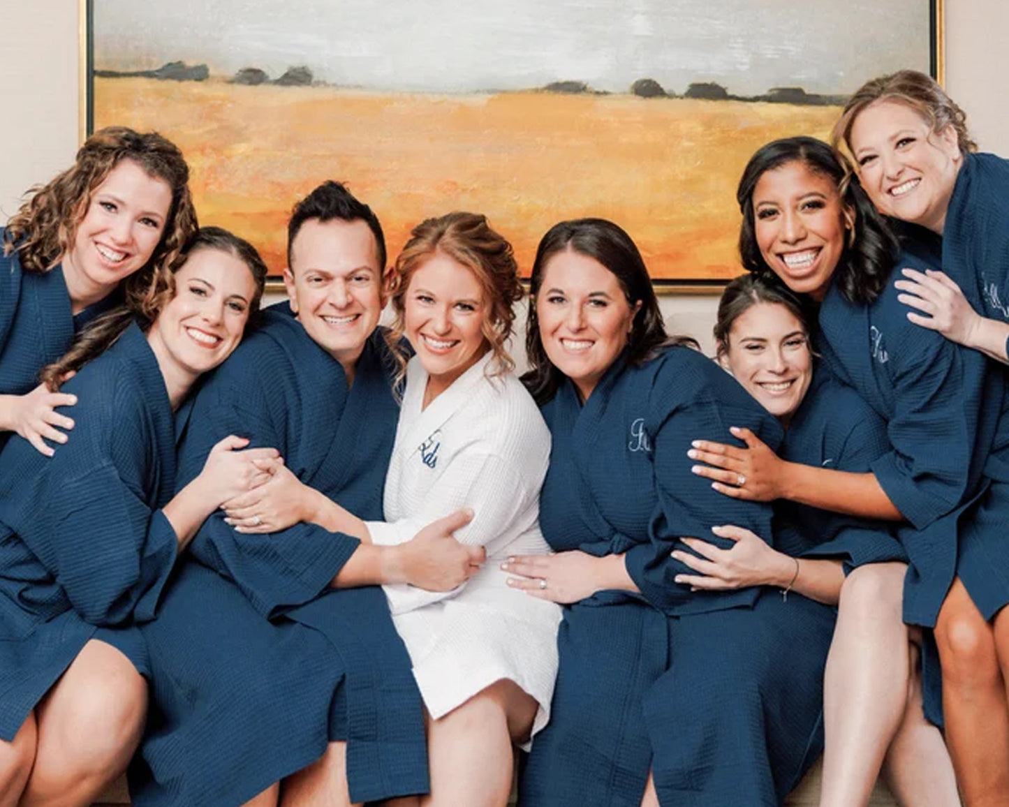 A joyful bridal party dressed in matching navy blue waffle robes, with the bride in white, all smiling and embracing each other in a warm, celebratory moment.