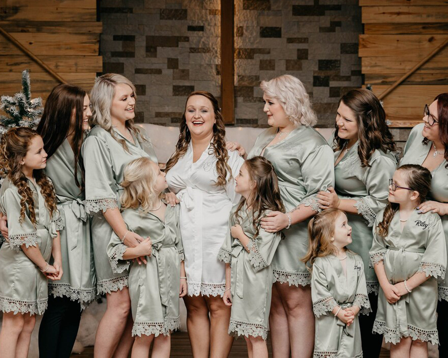 A bride in a white satin robe is surrounded by her bridesmaids and flower girls wearing matching sage green robes with lace trim, sharing smiles in a rustic indoor setting.