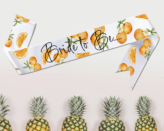A "Bride to Be" sash with an elegant orange-themed design on a white background, displayed above a row of pineapples.