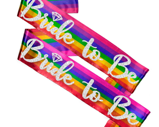A vibrant rainbow-colored sash with the words "Bride to Be" written in glittery silver script, featuring a diamond icon.