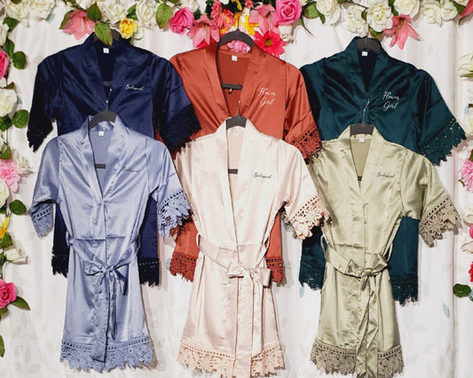 A collection of satin bridesmaid and flower girl robes in various colors, featuring delicate lace trim and embroidered text, displayed against a floral backdrop.