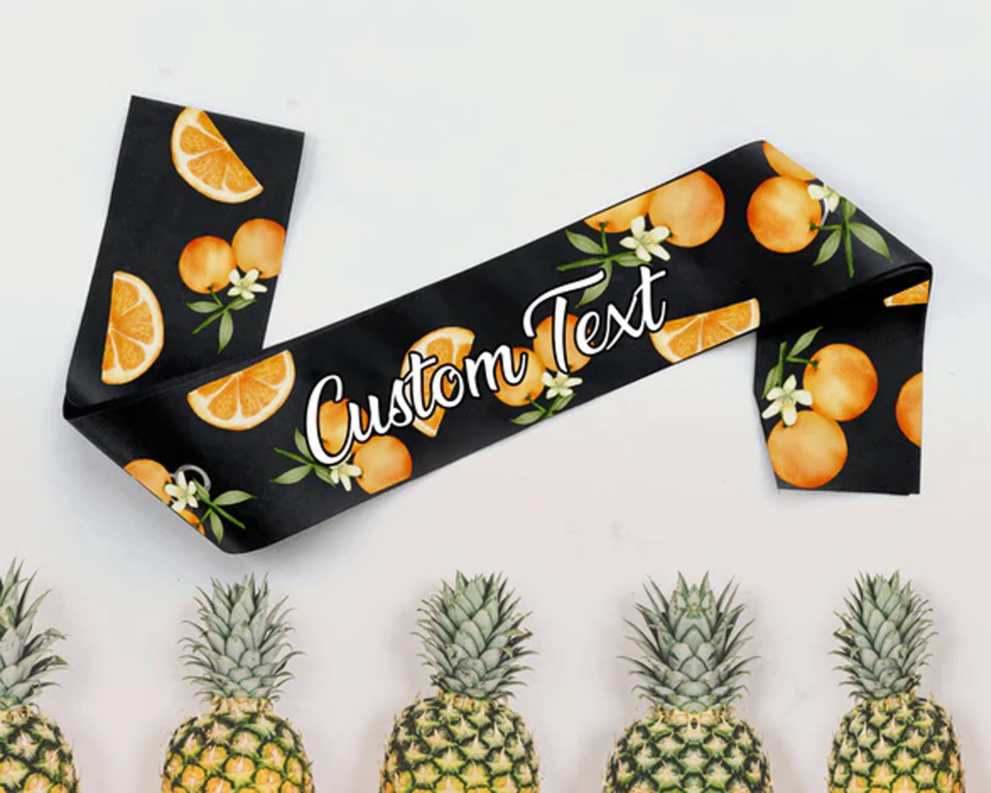 A black satin sash featuring vibrant orange slices and blossoms, customizable text, and a tropical pineapple backdrop.