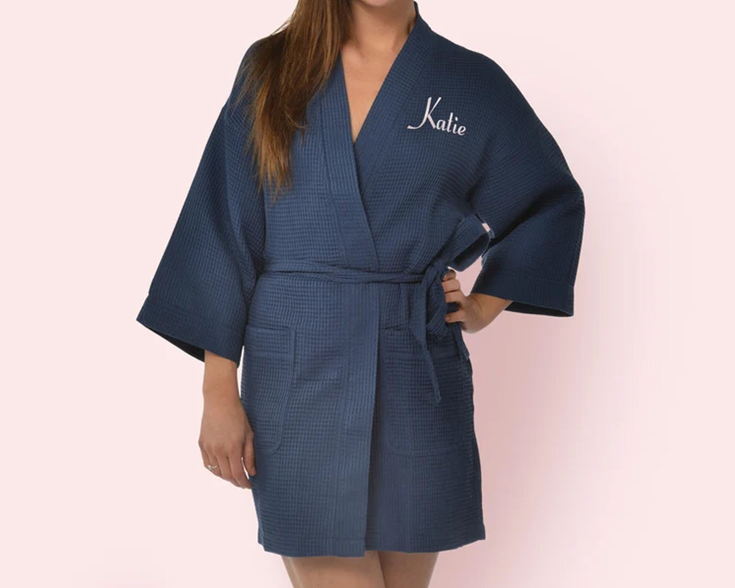 Woman wearing a navy blue waffle robe with personalized embroidery on the chest, standing against a soft pink background.