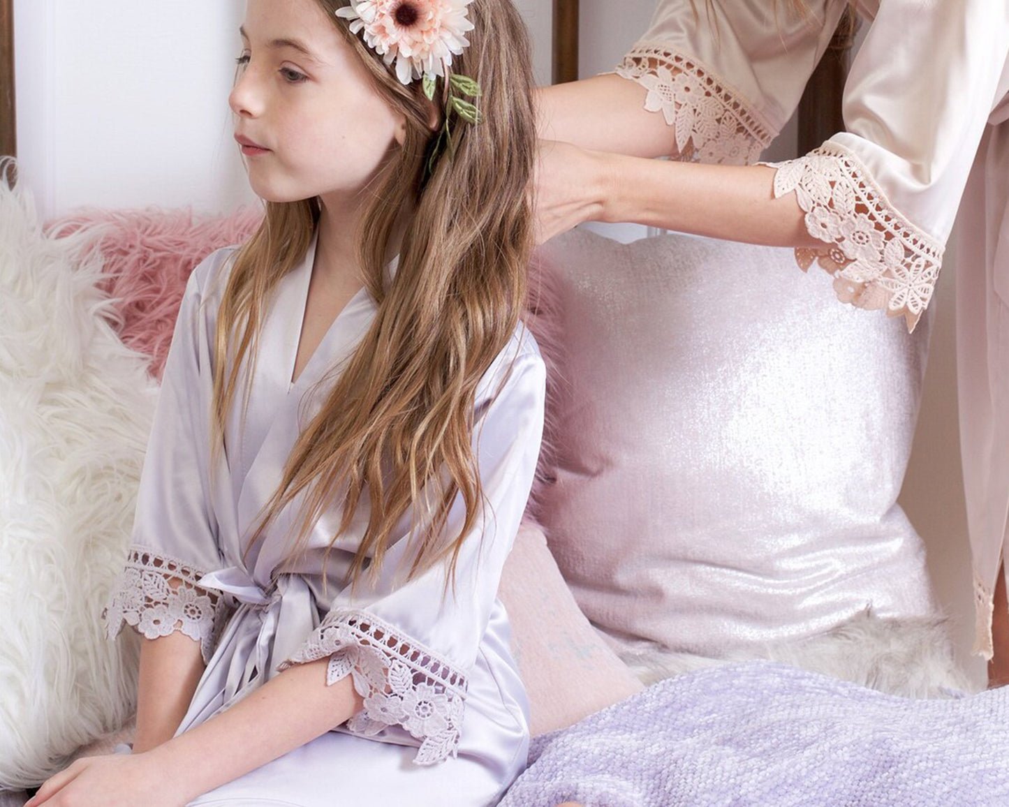 A young girl in a light lavender satin robe with lace trim sits on a cozy bed while an adult, also in a satin robe, styles her long brown hair adorned with a floral headband.