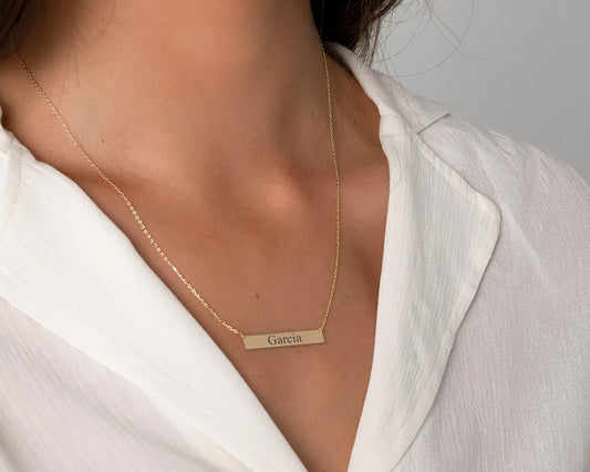 Elegant gold bar necklace featuring a personalized name engraving, worn with a white blouse.