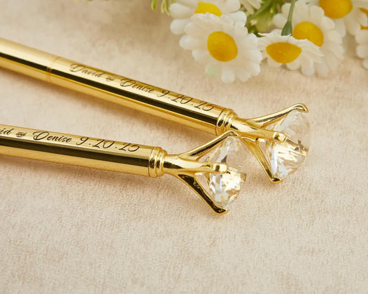 Elegant gold diamond-topped pens engraved with "David and Denise 9.20.25," placed on a soft beige surface with white and yellow flowers in the background.