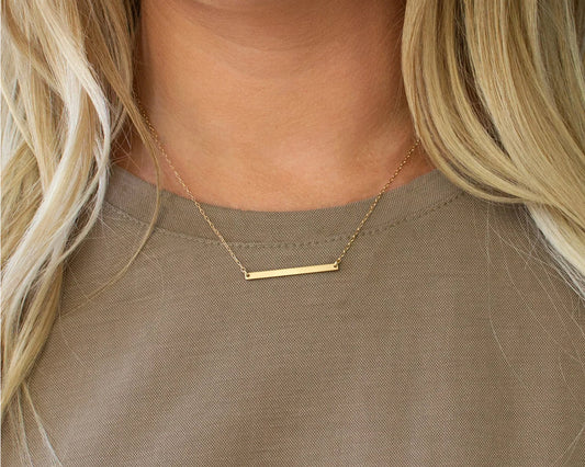 Elegant minimalist gold bar necklace on a delicate chain, worn by a woman in a neutral-toned outfit.