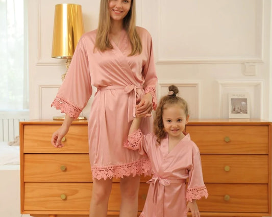 A mother and her young daughter wearing matching pink satin robes with lace trim, standing in a warmly lit bedroom, holding hands and smiling.