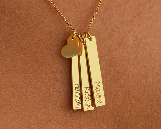 A close-up of a gold necklace featuring three vertical bar pendants engraved with the names "Hannah," "Robbie," and "Maxine," along with a small gold heart charm, worn against the skin.