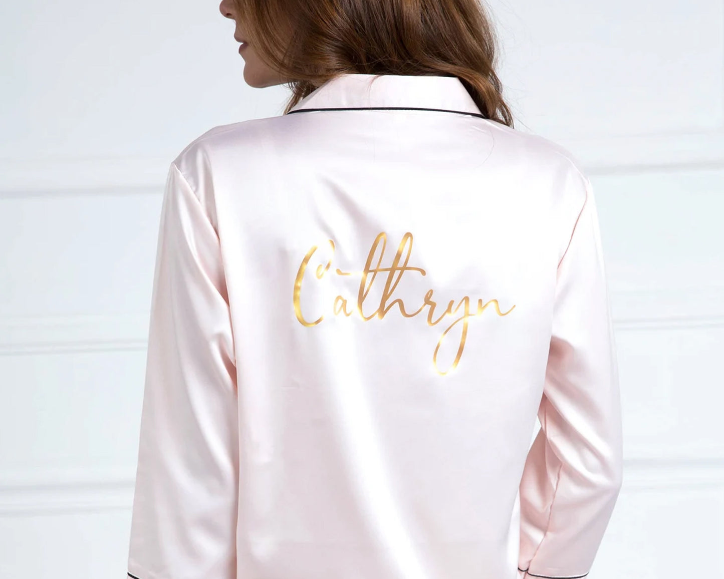 A woman wearing a blush pink satin pajama top with black piping, featuring a custom gold name print on the back.