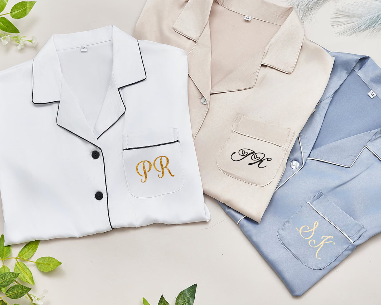 Three folded satin pajama tops in white, beige, and blue, each featuring embroidered initials on the pocket.
