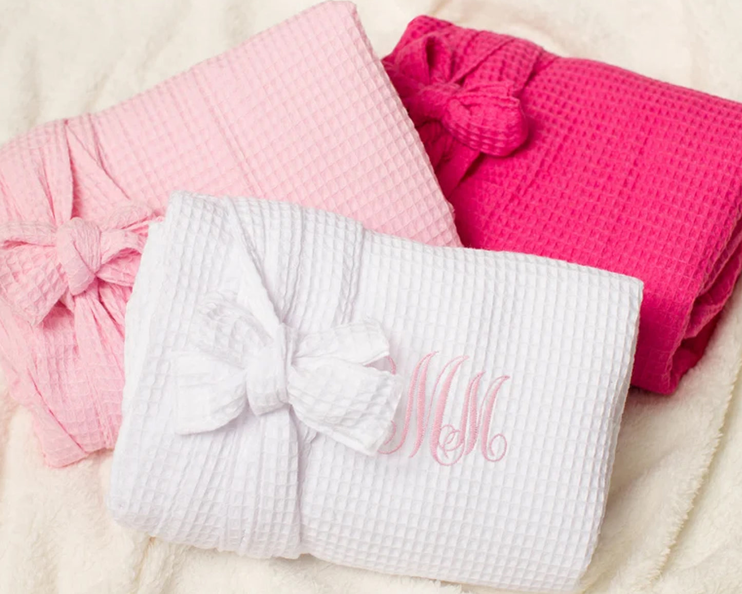 Three folded waffle-textured robes in white, light pink, and hot pink, neatly tied with bows, with the white robe featuring elegant monogram embroidery.