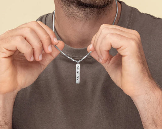 Man holding a silver bar pendant necklace engraved with Roman numerals, wearing a casual brown shirt.
