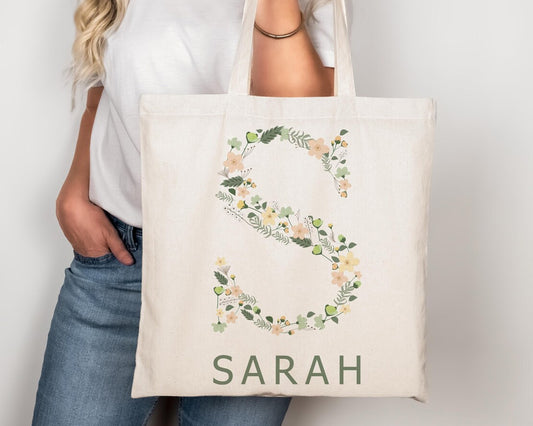 Beige tote bag featuring the letter "S" adorned with delicate green leaves and soft pastel flowers, with "SARAH" written below.