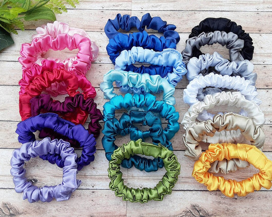 A collection of colorful satin scrunchies in shades of pink, blue, green, purple, yellow, white, gray, and black, arranged on a wooden surface with green leaves in the background.