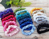 A variety of satin scrunchies in multiple vibrant colors, including pink, blue, green, purple, yellow, black, white, and gray, neatly arranged on a wooden surface with greenery in the background.