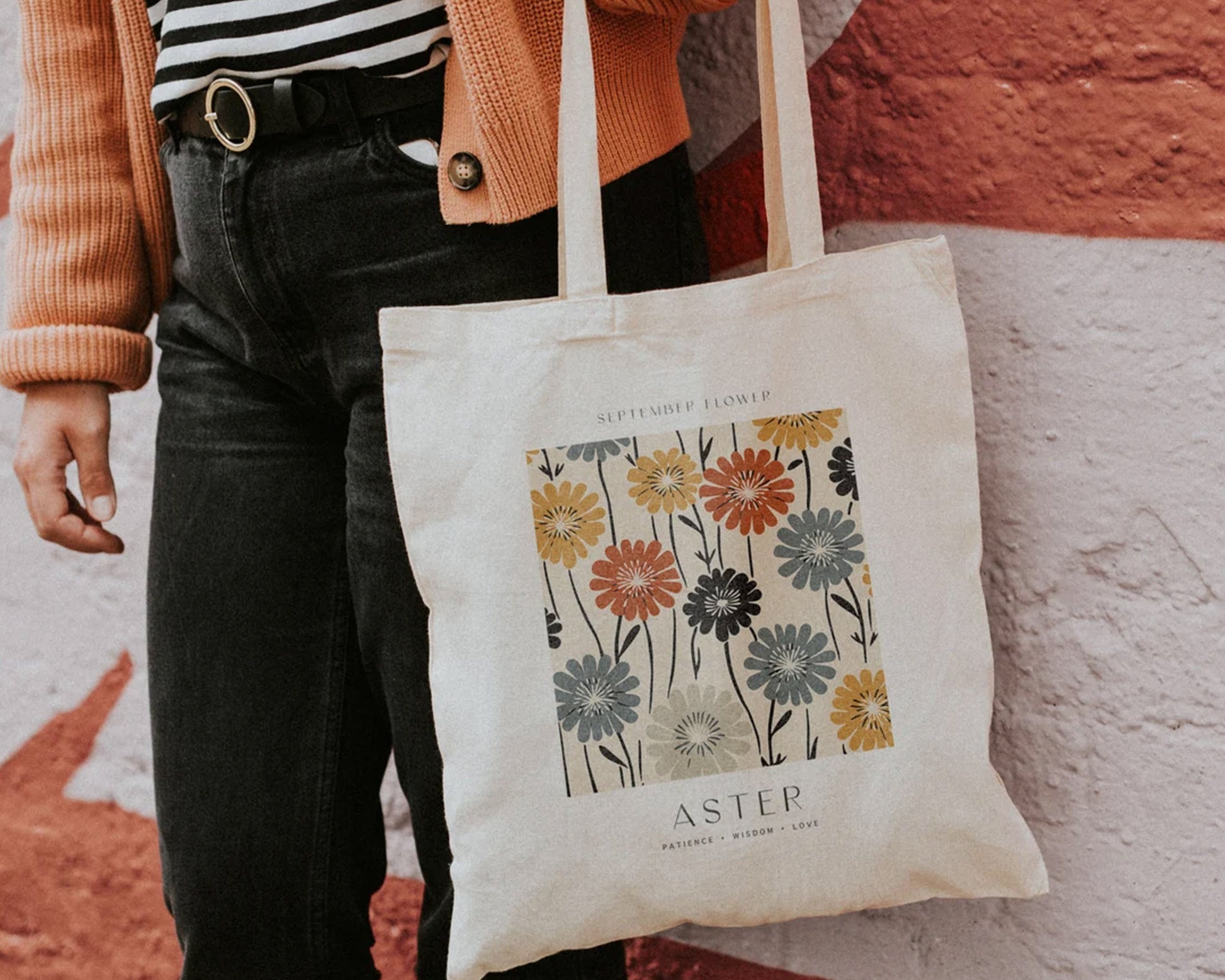 A beige canvas tote bag featuring a vintage-style floral design with asters in warm autumn colors, carried by a person wearing a striped shirt and cardigan.