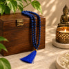 A royal blue beaded mala necklace with a matching tassel draped over a dark wooden box, styled with green leaves, a lit candle, and a small Buddha statue on a stone surface.