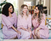Three women sitting together in matching blush pink robes with delicate lace trim, smiling and chatting.