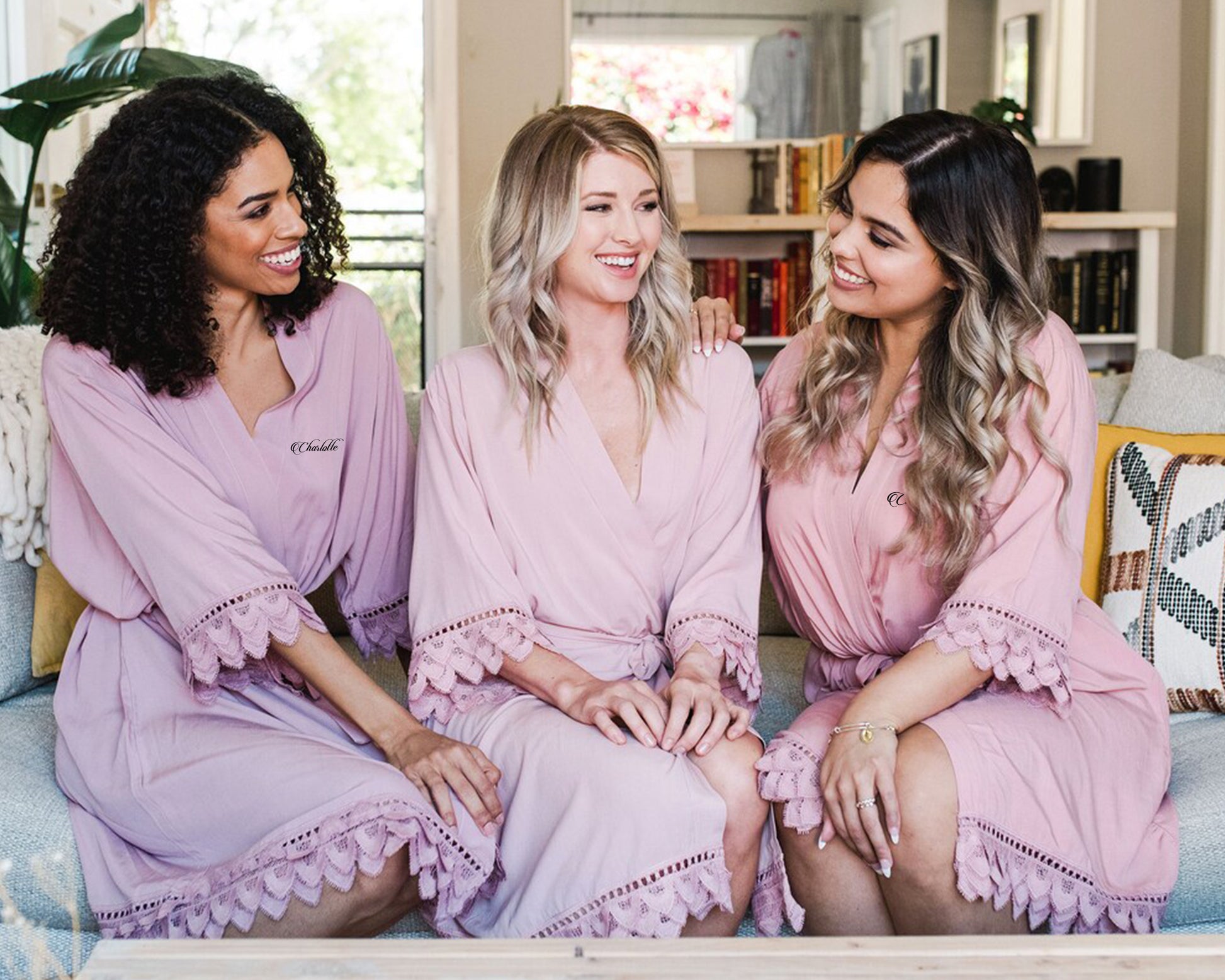 Three women sitting together in matching blush pink robes with delicate lace trim, smiling and chatting.