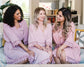 Three women sitting together in matching blush pink robes with delicate lace trim, smiling and chatting.
