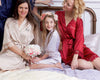 Bride, bridesmaid, and flower girl in satin robes sitting on a bed, smiling.
