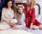 Bride, bridesmaid, and flower girl in satin robes sitting on a bed, smiling.