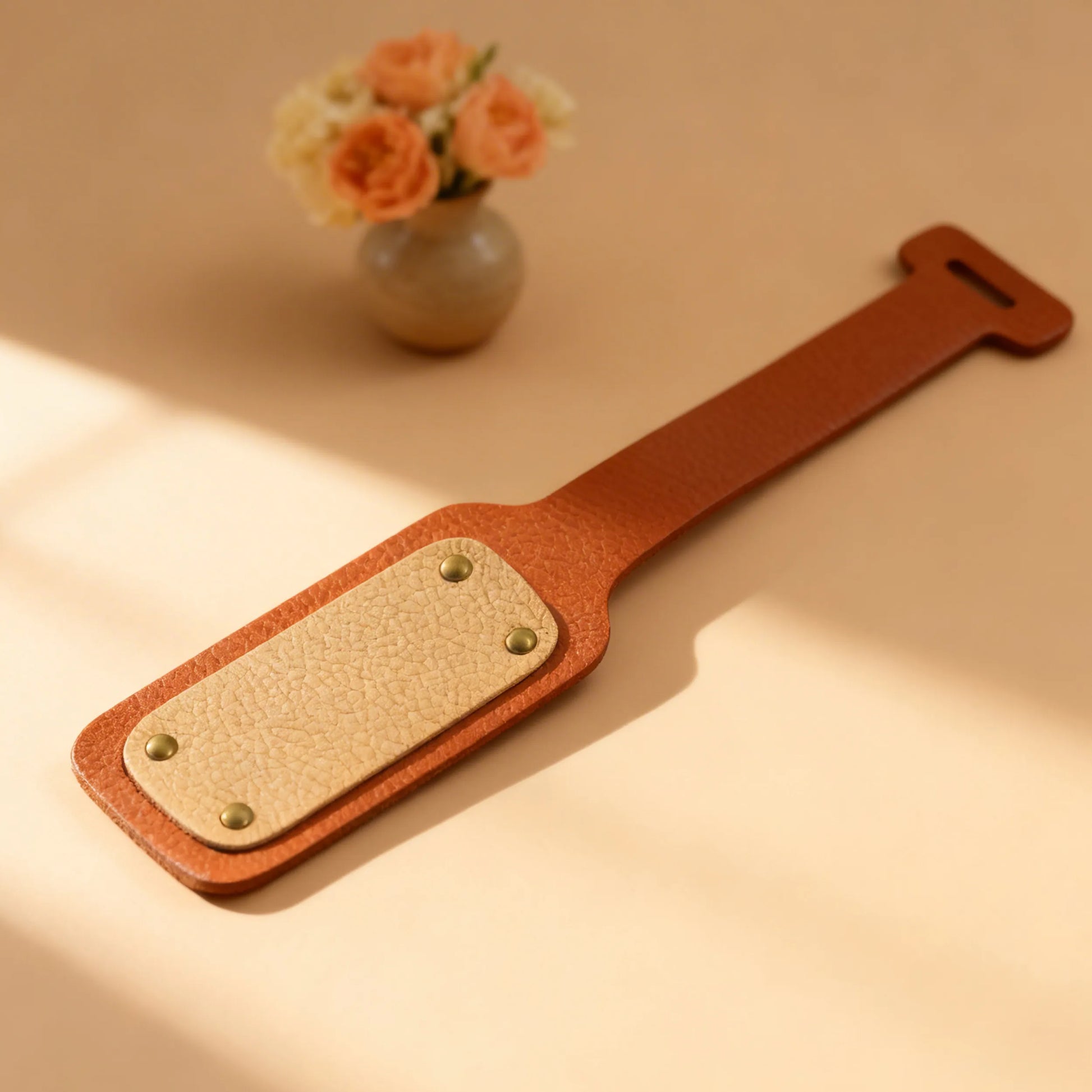 Brown leather strap with gold hardware on a beige surface with a vase of flowers in the background
