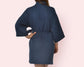 A woman wearing a navy blue waffle-textured robe, seen from the back, with a tied waist belt and wide sleeves, standing against a soft pink background.