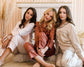 Three women sitting together in elegant satin pajama sets in soft neutral tones, featuring contrast piping and personalized embroidery.