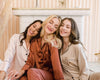 Three women sitting closely together, smiling and leaning on each other in soft satin pajama sets in champagne, blush, and copper tones, with delicate contrast piping.