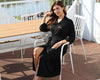 Woman in a black robe with "Bride" text, sitting outdoors on a patio chair.