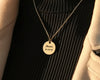 Gold round pendant necklace engraved with the name "Maria" and a date, worn over a black ribbed sweater with a beige coat.