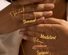  A collection of gold name necklaces with delicate cursive lettering, featuring names like Ophelia, Summer, Madeline, Olivia, and Simona, draped over a person's hands.