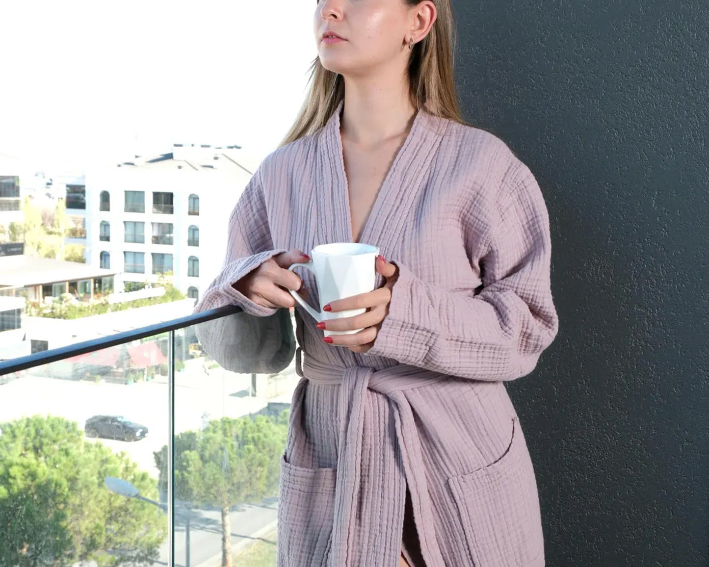 A woman wearing a mauve waffle-textured robe, holding a white coffee mug with red-painted nails. She is standing near a balcony with a blurred outdoor background.