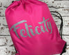 A pink drawstring bag with the name "Felicity" written in glittery silver cursive lettering, placed against a white brick background.