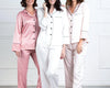Three women wearing luxurious satin full pajama sets in soft pastel shades, featuring button-up tops with contrast piping and matching pants.