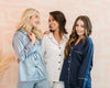 Three women in matching satin pajama sets—one in light blue, one in white, and one in navy—smiling and posing together against a soft pastel background.