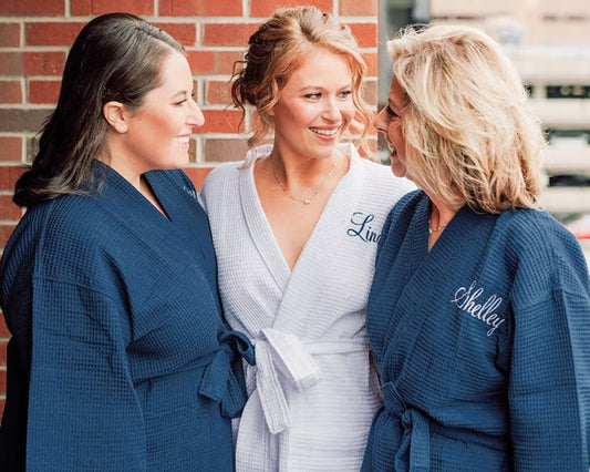 Three women wearing personalized waffle robes, two in navy and one in white, smiling together against a brick wall.