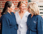 Three women wearing personalized waffle robes, two in navy and one in white, smiling together against a brick wall.