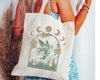 A beige canvas tote bag featuring a mystical fairy design with mushrooms, sunflowers, and crescent moons, carried by a woman wearing a floral dress.