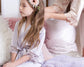 A young girl in a light lavender satin robe with lace trim sits on a cozy bed while an adult, also in a satin robe, styles her long brown hair adorned with a floral headband.