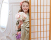A young girl in a sage green satin robe with lace trim, holding a bouquet of pink and white flowers, smiling while peeking from behind a wooden room divider.
