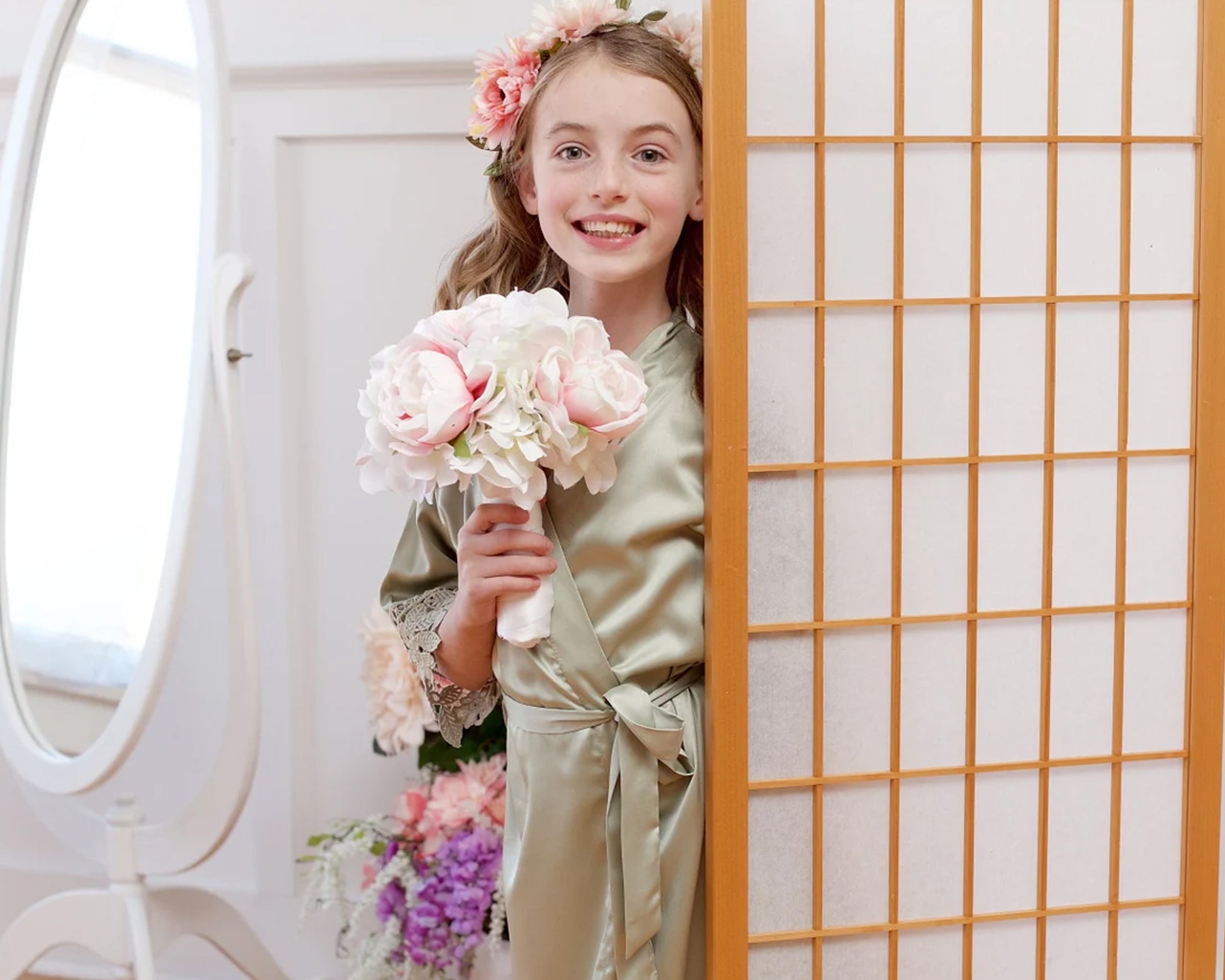 A young girl in a sage green satin robe with lace trim, holding a bouquet of pink and white flowers, smiling while peeking from behind a wooden room divider.