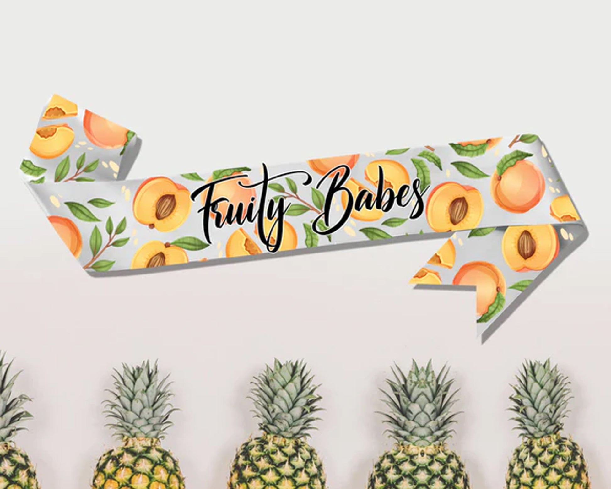 A "Fruity Babes" sash with a peach-themed design on a light gray background, displayed above a row of pineapples.