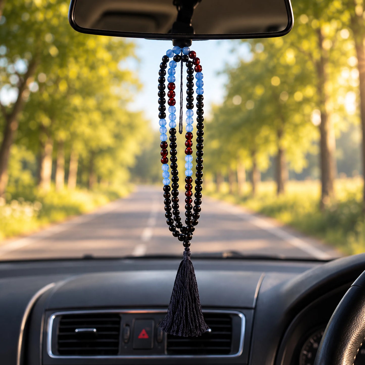 A beaded car hanging ornament with a tassel is suspended from a rearview mirror inside a car, with a blurred tree-lined road visible through the windshield.
