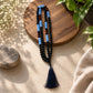 A beaded necklace with black, light blue, and dark brown beads, featuring a navy blue tassel, styled on a round wooden board with a stone and green leaves.
