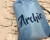 A light blue drawstring bag with the name "Archie" written in dark blue glittery cursive lettering, placed against a rustic white brick background.