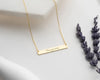 Elegant gold bar necklace with "Love you more" engraving, displayed on a white surface with lavender sprigs.
